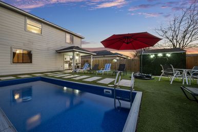 Suburban two-story home backyard at sunset with a rectangular blue pool, metal ladder, stepping-stone path across artificial turf, lounge chairs and a large red patio umbrella next to a wooden fence and evening sky.