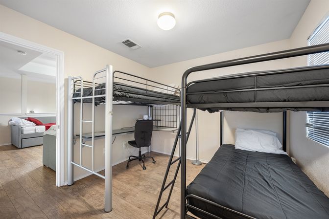 Cozy, compact apartment bedroom with two metal loft bunk beds in black bedding, built-in study desk with swivel chair, wood-look floors, and a bright window.