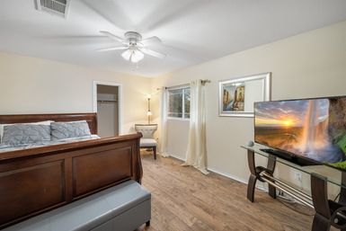 Cozy master bedroom with dark wooden sleigh bed and gray bedding, tufted bench, corner armchair and floor lamp, ceiling fan, window with sheer curtains, hardwood-look floors, and a flat-screen TV on a glass stand displaying a waterfall sunset.