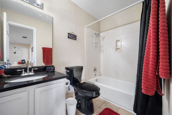 Residential bathroom with black toilet and black granite countertop, white vanity and tub-shower, red towels and rug, beige tile floor and large wall mirror — clean contemporary styling with pops of red.