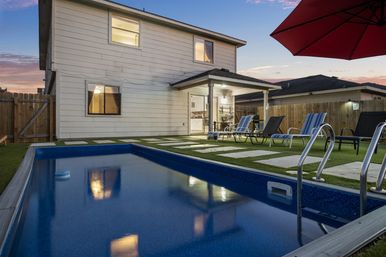 Inviting backyard rectangular pool at dusk beside a two-story suburban house with covered patio, lounge chairs, red umbrella, stepping-stone lawn, and wooden fence.