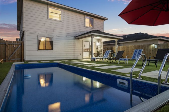 Inviting backyard rectangular pool at dusk beside a two-story suburban house with covered patio, lounge chairs, red umbrella, stepping-stone lawn, and wooden fence.