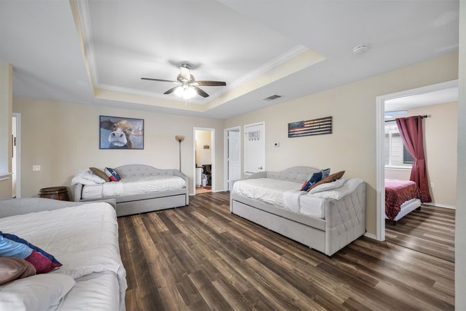 Bright, spacious guest bedroom with three tufted twin daybeds, dark wood-look floors, tray ceiling with ceiling fan, whimsical cow and American flag wall art, floor lamp, and doorway to adjacent room with red curtains — family-friendly rental sleeping area.