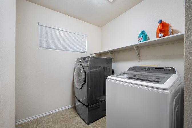 Compact home laundry room with gray front-load washer and white top-load washing machine on beige tile, wall shelf holding detergent bottles and a small blind-covered window.