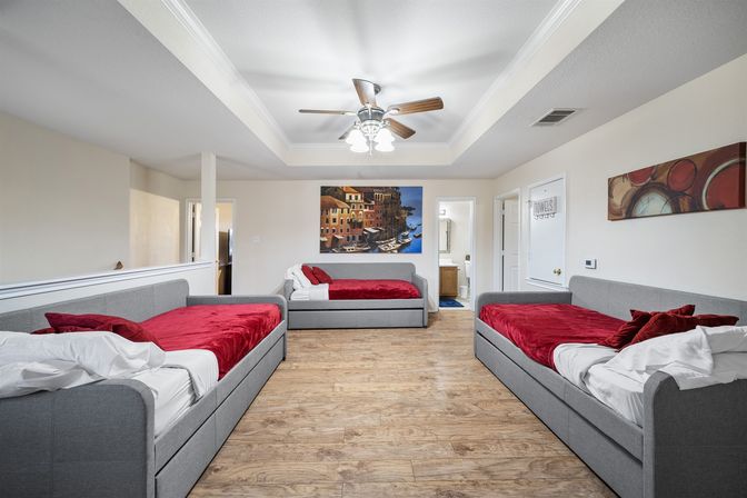 Spacious vacation rental guest room with three gray daybeds dressed in red blankets and pillows, wood-look flooring, tray ceiling with ceiling fan, coastal harbor wall art, and a doorway to an en suite bathroom — modern family suite interior.