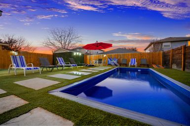 Inviting backyard swimming pool at sunset with deep-blue water, striped lounge chairs, red patio umbrella, stepping stones on turf and wooden fence