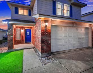 Twilight view of a two-story suburban brick-and-siding home with a bright blue front door, illuminated covered porch, white two-car garage, concrete driveway, rock landscaping, and a patch of green lawn.