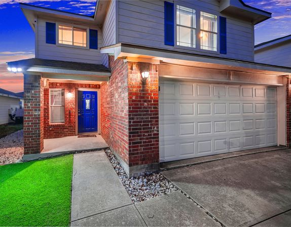 Twilight view of a two-story suburban brick-and-siding home with a bright blue front door, illuminated covered porch, white two-car garage, concrete driveway, rock landscaping, and a patch of green lawn.