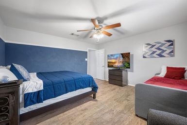 Spacious modern bedroom with navy accent wall, two beds (navy comforter and red throw), ceiling fan, TV on a dresser, wood-look floors and geometric wall art