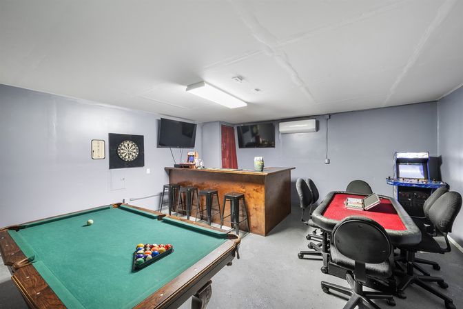 Basement entertainment room with green pool table and racked balls, rustic bar with stools, poker table, dartboard, arcade cabinet and wall-mounted TVs