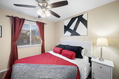 Cozy modern bedroom interior with red bedding and round bolster pillows, black accent pillows, abstract black-and-white wall art, red curtains, ceiling fan, and white nightstand with lamp.