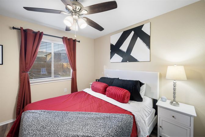 Cozy modern bedroom interior with red bedding and round bolster pillows, black accent pillows, abstract black-and-white wall art, red curtains, ceiling fan, and white nightstand with lamp.