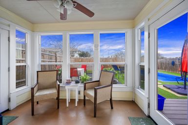 Bright enclosed sunroom with two beige armchairs and a white side table under a ceiling fan, windows framing a fenced suburban backyard with colorful patio chairs and a blue swimming pool.