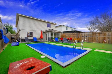 Suburban two-story house backyard with bright blue rectangular pool and ladder, green artificial turf, lounge chairs and red patio umbrella, cornhole boards and a small putting green behind a wooden privacy fence.