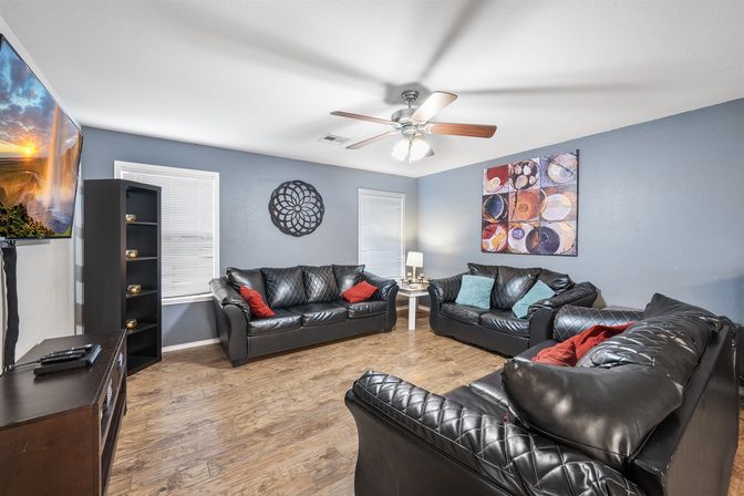 Cozy modern living room interior with three black leather sofas on wood-look flooring, blue-gray walls, ceiling fan, colorful abstract wall art, red and teal throw pillows, shelving and wall-mounted flat-screen TV.