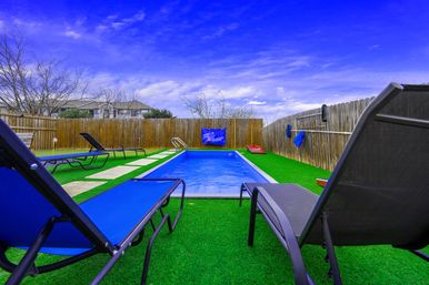 Rectangular backyard pool surrounded by vibrant green artificial turf and a wooden privacy fence, with blue lounge chairs, a hanging pool float, cornhole board, and a bright blue sky