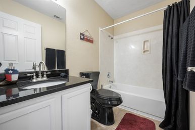 Small modern bathroom with white vanity and black granite countertop, chrome faucet, black toilet and black shower curtain around a white bathtub, beige walls, red bath mat and a small patriotic star Welcome plaque.
