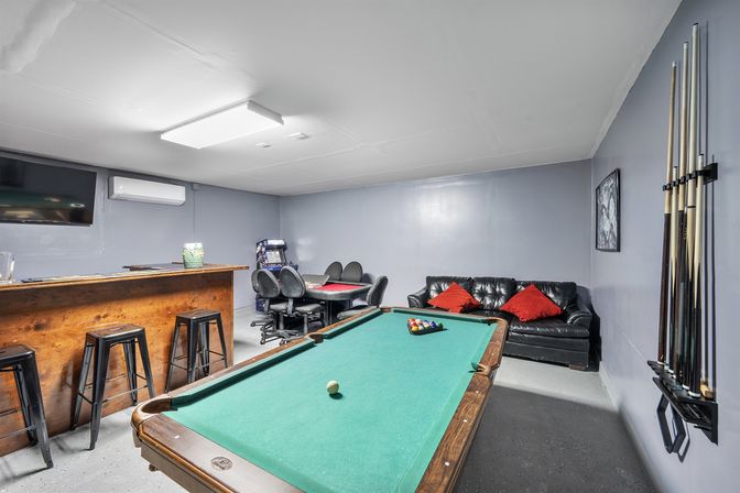 Basement game room with a green-felt pool table and wall cue rack, wooden bar with stools, poker table with swivel chairs, black leather couch with red pillows, TV and arcade-style game.