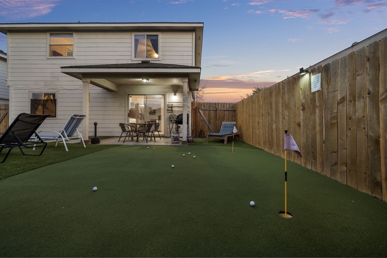 Suburban backyard at sunset featuring an artificial putting green with golf balls and flags, a covered patio with a dining table and chairs attached to a two-story white house, wooden privacy fence and lounge chairs.