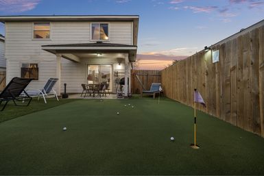 Suburban backyard at sunset featuring an artificial putting green with golf balls and flags, a covered patio with a dining table and chairs attached to a two-story white house, wooden privacy fence and lounge chairs.