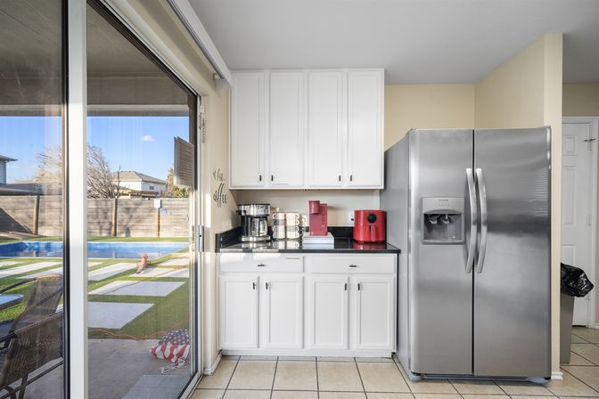 Bright modern kitchen with white cabinets and black countertop holding a stainless coffee maker and red espresso machine, stainless steel side-by-side refrigerator, tiled floor and sliding glass door opening to a suburban backyard with pool and patio