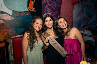 Three smiling friends at a lively bar celebrating a birthday — center woman wears a tiara and glittery sash, flanked by women in green and pink dresses.