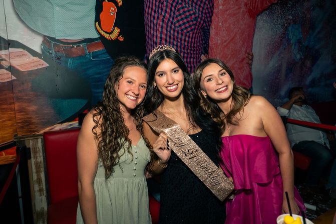 Three smiling friends at a lively bar celebrating a birthday — center woman wears a tiara and glittery sash, flanked by women in green and pink dresses.