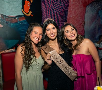 Three smiling friends at a lively bar celebrating a birthday — center woman wears a tiara and glittery sash, flanked by women in green and pink dresses.