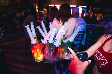 Glow-lit tray of novelty syringe shots filled with colorful liquids being offered in a crowded bar, lively nightlife party scene
