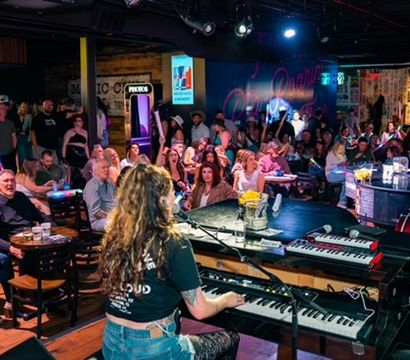 Keyboardist on stage playing to a packed live music bar, energetic crowd at tables cheering under colorful stage lights, neon sign and disco ball overhead — lively nightlife scene.