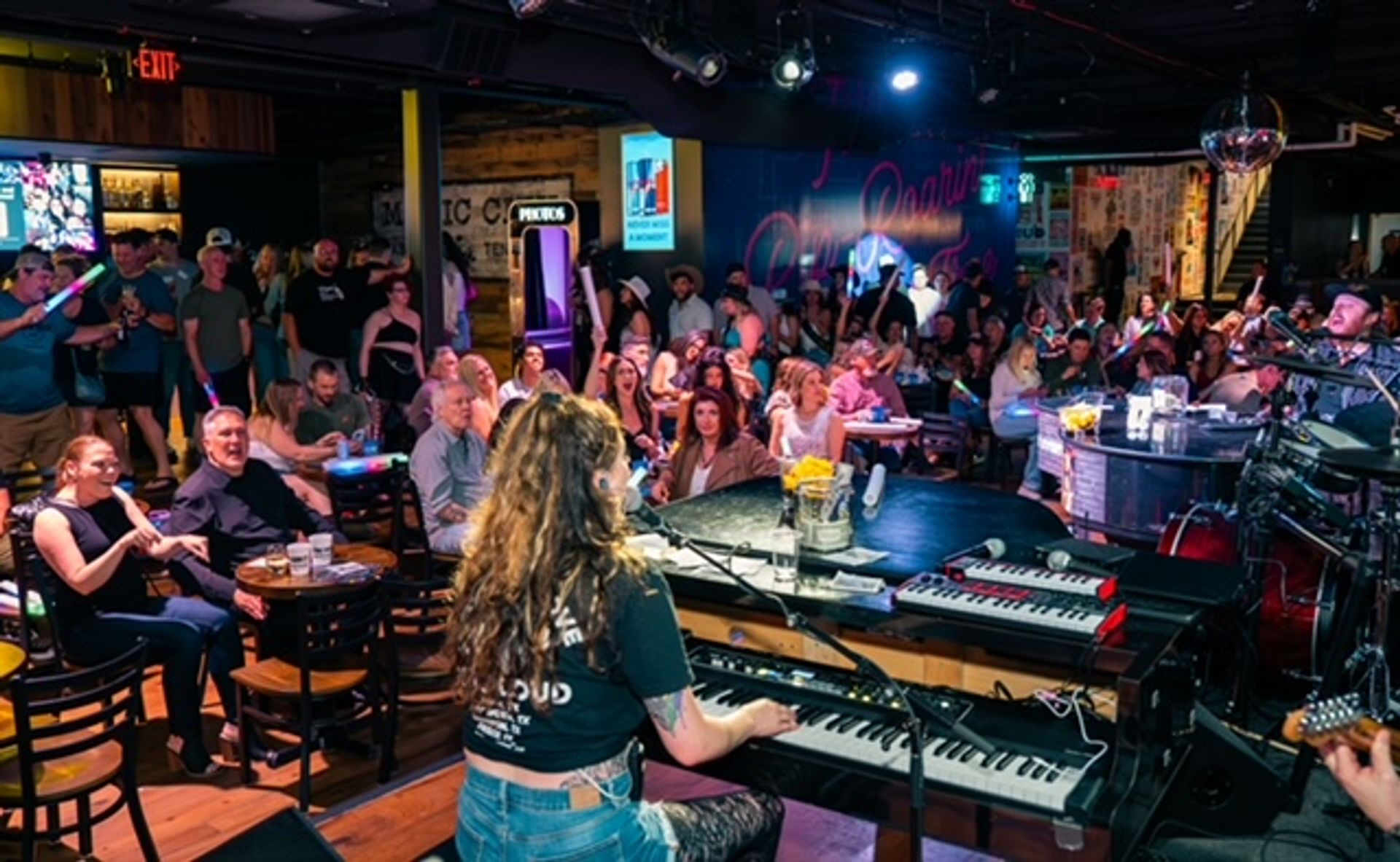 Keyboardist on stage playing to a packed live music bar, energetic crowd at tables cheering under colorful stage lights, neon sign and disco ball overhead — lively nightlife scene.