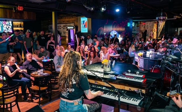 Keyboardist on stage playing to a packed live music bar, energetic crowd at tables cheering under colorful stage lights, neon sign and disco ball overhead — lively nightlife scene.