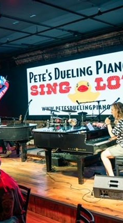 Dueling piano bar with two grand pianos and performers singing into microphones on stage, lively seated audience, colorful stage lighting and a large illuminated backdrop reading 'SING LOUD'.