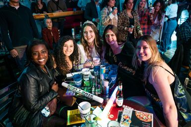 Five friends smiling around a crowded bar table under colorful neon lights, with beer, cups, snacks and party props; one wearing a tiara and sash for a lively nightlife celebration.