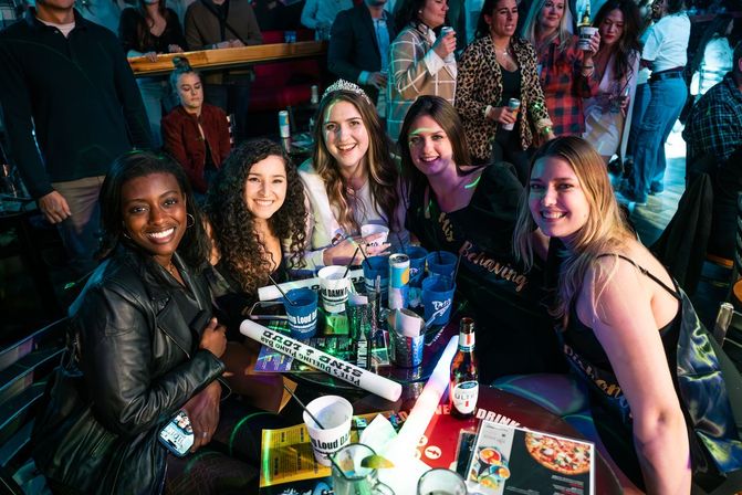 Five friends smiling around a crowded bar table under colorful neon lights, with beer, cups, snacks and party props; one wearing a tiara and sash for a lively nightlife celebration.