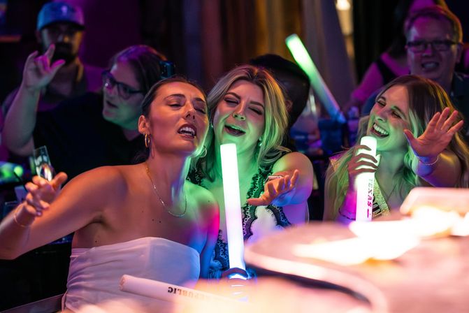 Three friends at a lively nightclub bar laughing and singing while holding glowing LED foam sticks under colorful party lights — upbeat nightlife celebration.