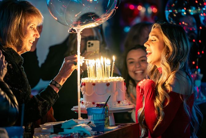 Young woman in a red dress smiling as an older woman lights many candles on a decorated birthday cake at a festive indoor party with balloons and colorful lights.