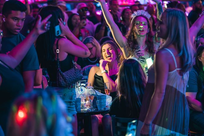Neon-lit bar crowd at night — lively bachelorette party with a woman wearing a bride sash and tiara cheering as friends dance and toast with champagne