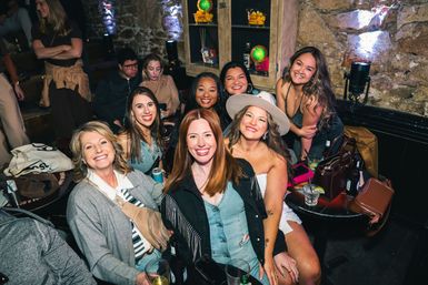 Smiling group of women at a cozy rustic stone-walled bar, gathered around a small table with cocktails, beer bottles, handbags and colorful mood lighting — lively downtown nightlife scene.