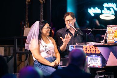 Bachelorette in veil and sash laughing as a pianist-singer performs at a neon-lit piano bar with guitars on stage and a lively audience.