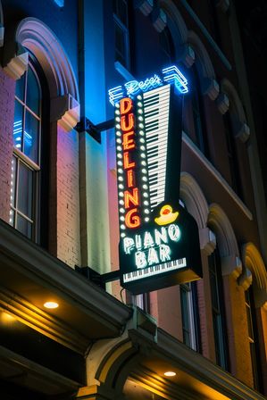 Neon 'Dueling Piano Bar' sign shaped like piano keys with a rubber duck on a historic downtown brick storefront at night