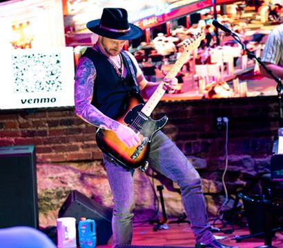 Tattooed guitarist in a fedora playing electric guitar on a colorful indoor bar stage with tables in the foreground and a QR payment sign, lively nightlife vibe.