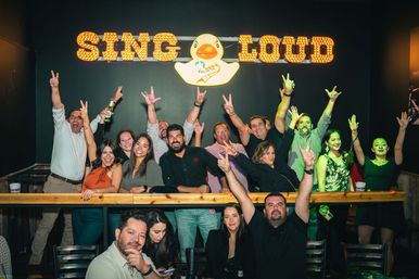Energetic group of adults cheering and flashing peace signs at an indoor karaoke bar, gathered behind a wooden railing under a lit "SING LOUD" sign with a rubber duck logo and colorful stage lighting.