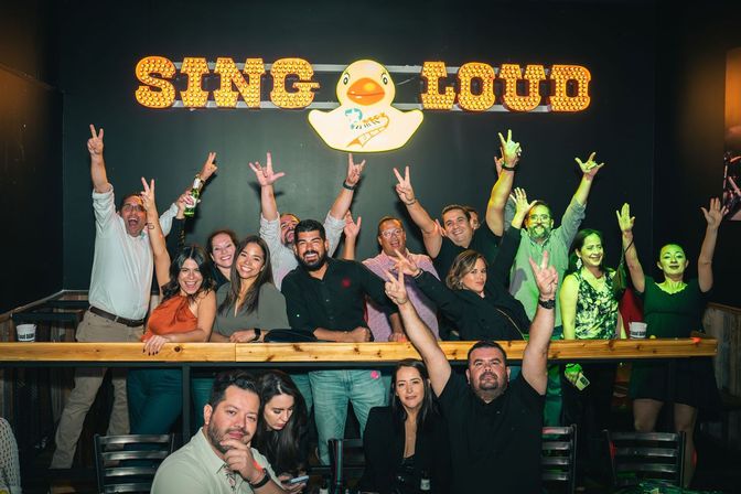 Energetic group of adults cheering and flashing peace signs at an indoor karaoke bar, gathered behind a wooden railing under a lit "SING LOUD" sign with a rubber duck logo and colorful stage lighting.