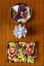 Overhead view of two gourmet platters on a warm wooden table: a round charcuterie tray with brie, salami, olives, grapes, crackers and jam, and a rectangular tropical fruit tray with watermelon, dragon fruit, pineapple, starfruit, grapes and assorted berries, separated by a white coral centerpiece.