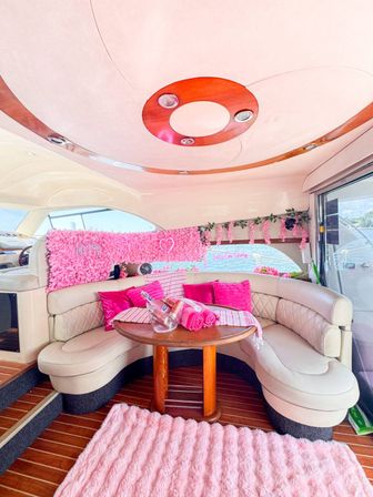 Pink-themed luxury yacht interior with curved cream leather banquette, round wooden table holding rolled pink towels and ice bucket, pink shag rug, floral wall panel and marina view through windows