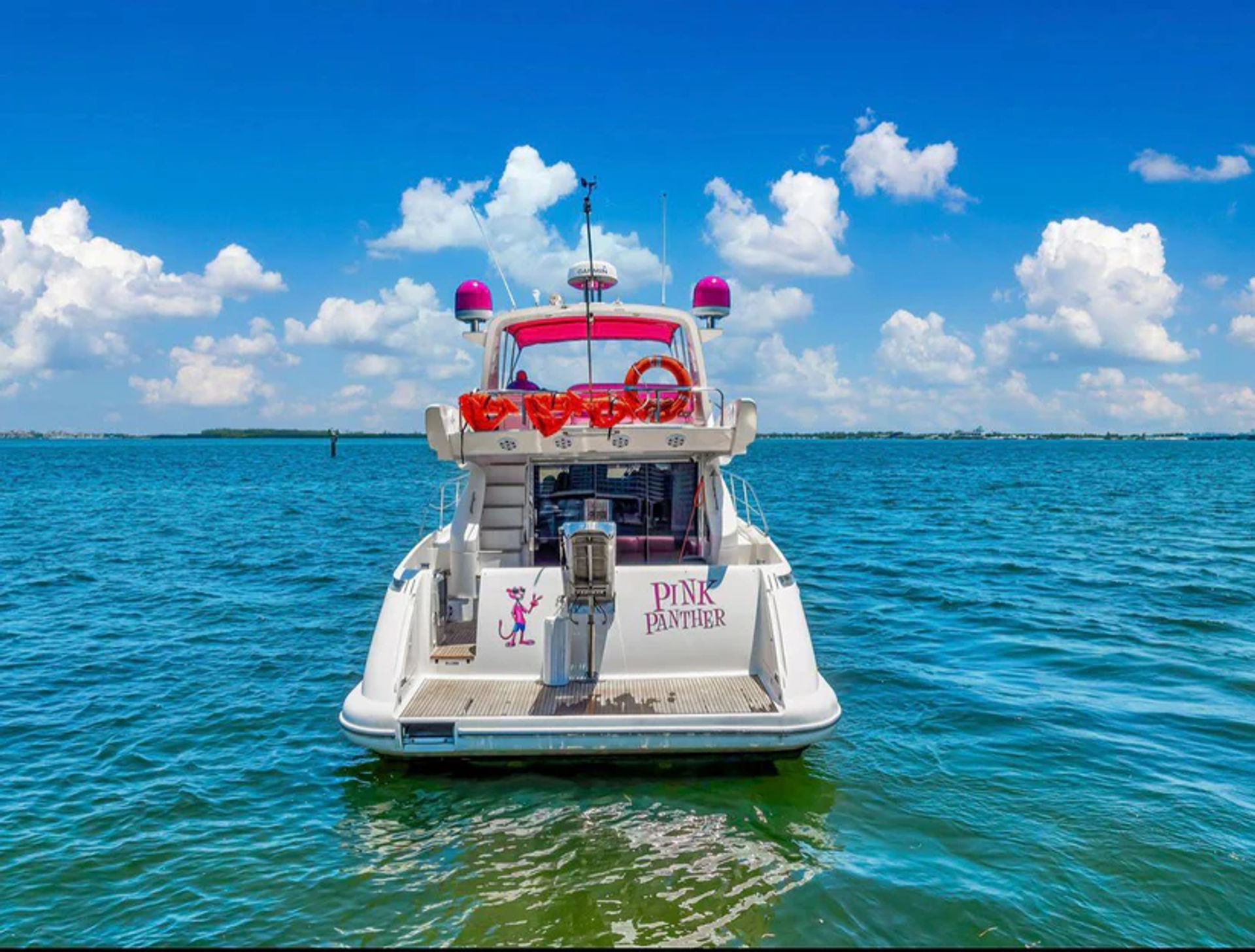 White motor yacht with pink accents and orange life rings floating on turquoise coastal water under a bright blue sky with fluffy clouds