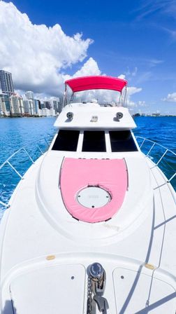 White luxury yacht with pink canopy and matching sunpad cruising turquoise bay with a coastal downtown skyline under bright blue sky and fluffy clouds