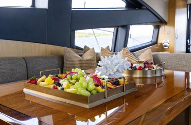 Fresh fruit tray with grapes, pineapple and berries plus decorative white coral on a glossy wooden dining table inside a luxury yacht cabin with cushioned seating and a water view.