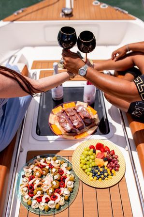 Two people clinking black wine glasses on a yacht deck, toasting over charcuterie, shrimp-and-olive appetizers and a colorful fruit platter on teak decking.